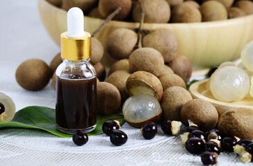 A bunch of ripe longan fruits lay on the ground, and in a wooden bowl, a glass bottle with a squeeze cap containing a dark brown liquid lay on top of the green leaves, next to it were black seeds.


