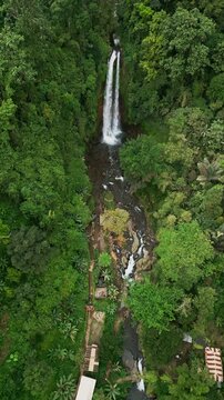 High above Bali&rsquo;s Gitgit Waterfall, a vertical drone captures the powerful stream falling into a rocky river, wrapped in lush green jungle and vibrant tropical vegetation.