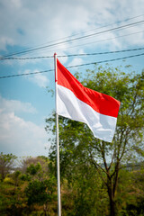 Red and white Indonesian flag waving on pole against blue sky and green trees outdoors