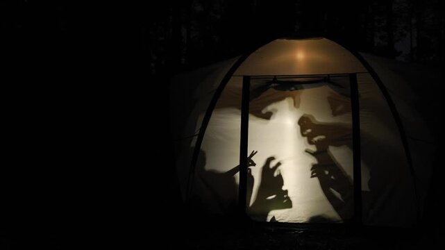 Silhouettes of children create playful shadow puppets inside a glowing tent during nighttime in the forest, capturing a fun and creative moment of outdoor adventure