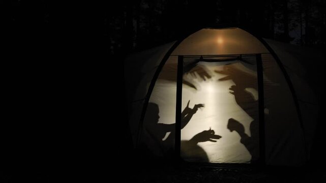 Silhouettes of children create playful shadow puppets inside a glowing tent during nighttime in the forest, capturing a fun and creative moment of outdoor adventure