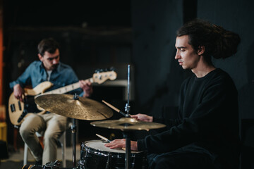 Two young musicians practicing together in a studio, one playing the drums and the other playing...