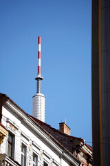 Obraz premium Tall communication tower with red and white stripes rises above rooftops against a clear blue sky. Architectural details of the building roof and chimney are visible. Clear daytime urban scene.