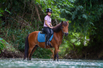 Young person rides brown horse through shallow river, surrounded by lush greenery, wearing helmet and equestrian gear