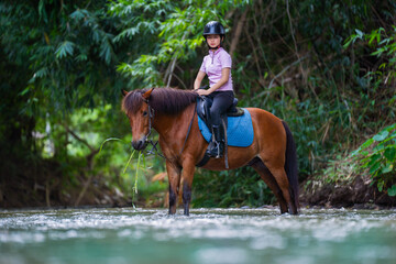 Person riding brown horse through shallow river, surrounded by lush greenery, wearing helmet and casual attire