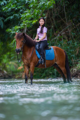 Young woman rides brown horse through shallow river, surrounded by lush greenery, creating serene and adventurous atmosphere