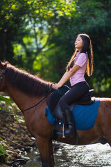 Young woman joyfully rides horse through forest stream, surrounded by lush greenery and sunlight filtering through trees