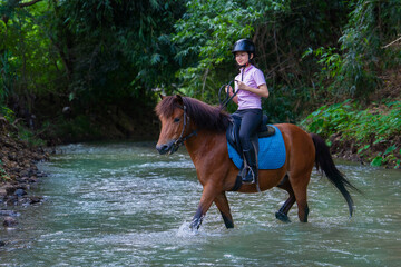 Person riding brown horse through shallow forest stream, wearing helmet and casual attire, enjoying peaceful nature experience