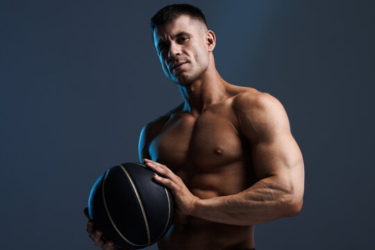 Muscular model sports man with a basketball on dark background. Fashion portrait of strong brutal guy. Male flexing his muscles.
