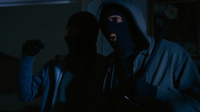 Two unrecognizable thieves in black clothes and balaclavas standing together in living room and discussing plan of stealing in middle of night