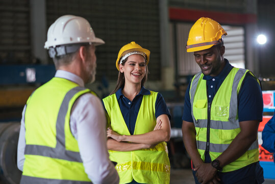 Diverse team of industrial workers wearing safety helmets and reflective vests receiving instructions from supervisor in manufacturing factory, focusing on teamwork, safety, and productivity.