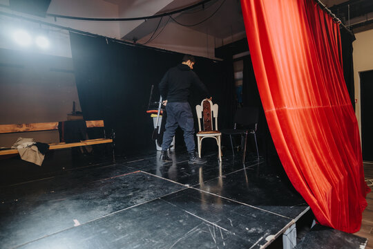 The image shows a scene where a stagehand is preparing a chair among props on a dimly lit theater stage with red curtains.