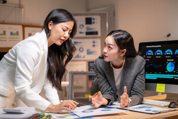 Two women are sitting at a desk with a computer monitor in front of them