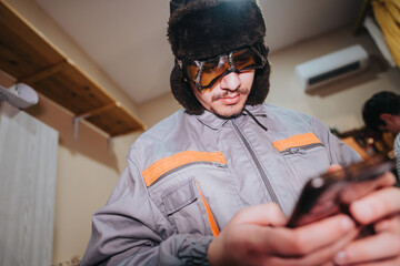Focused man in outdoor attire, including fur hat and goggles, looking at a smart phone in a...
