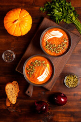 Overhead view of two bowls of creamy pumpkin soup garnished with cream and pumpkin seeds, surrounded by a whole pumpkin, fresh parsley, red onions, bread slices. Homegrown organic eco food 