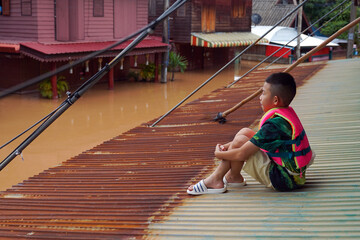 Asian boy wearing a life jacket sits hugging his knees on a tin roof, unable to go out because his house is flooded.