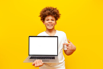 cheerful african american boy in white t-shirt showing blank laptop screen on yellow isolated...