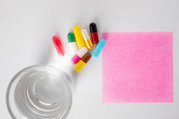 Top view of glass of water, many pill capsules and pink note paper on white background with copy space.