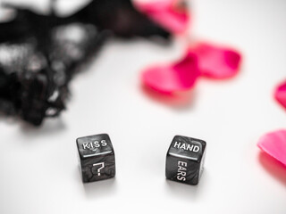Two black romantic dice with words and symbols, surrounded by lace lingerie and pink rose petals. Dice with the words “Kiss” and “Hand” written on them.