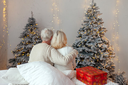elderly senior couple in sweaters celebrate new year and hug at home, old family grandma and grandpa look at garland and decorated christmas tree