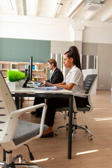 African American woman and European young man in business suit sitting in office and typing on...