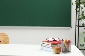 Different school stationery, glasses and apple on white table near blackboard indoors, space for...