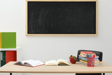 Different stationery and red apple on wooden table near blackboard indoors