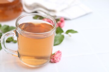 Aromatic tea in glass cup, roses and green leaves on white table, closeup. Space for text
