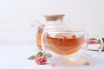 Aromatic tea in glass cup, teapot, roses and green leaves on light table, closeup. Space for text