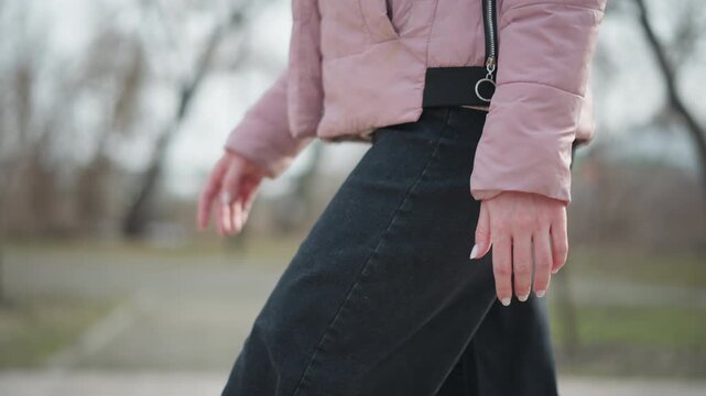 Close-up of female hand relaxed by side while walking in cold weather, wearing pink winter jacket and black pants, soft natural lighting, blurred outdoor background, focus on casual motion