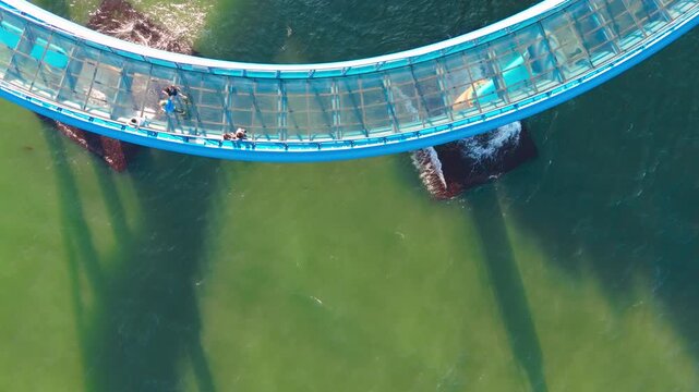 Aerial view of concrete bridge supports and foundations submerged in clear, green ocean water. Shadows stretch across the water surface.
