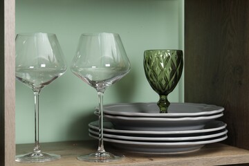 Different plates and glasses on wooden shelf near green wall indoors, closeup