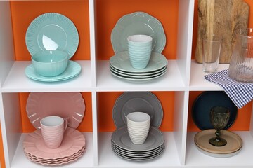 Shelving unit with different dishware, cups and glasses near orange wall indoors