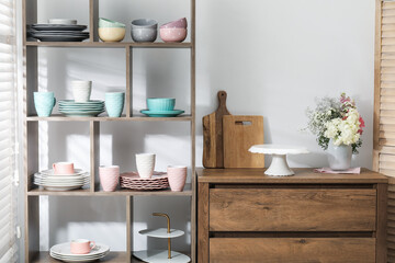 Shelving unit with different dishware, cups and floral decor near white wall indoors