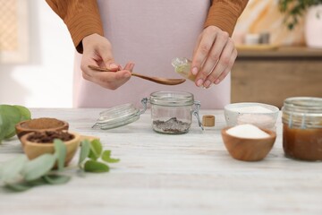 Making natural scrub. Woman adding essential oil into jar at white wooden table, closeup