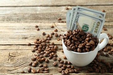 Dollar banknotes and coffee beans in cup on wooden table, closeup. Space for text