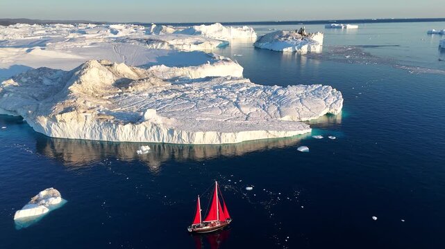 Little red sailboat cruising among floating icebergs in Disko Bay glacier during midnight sun season of polar summer. Ilulissat, Greenland. Global warming and melting glaciers.