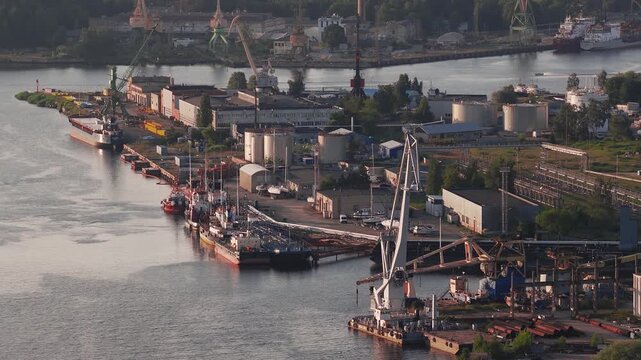 A shipyard in Riga, Latvia, with cranes in motion, docked ships, and industrial buildings near the Daugava River during golden hour.