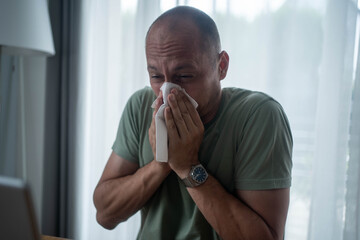 Sick man blowing nose while working from home on laptop, allergy or flu symptoms in home office...