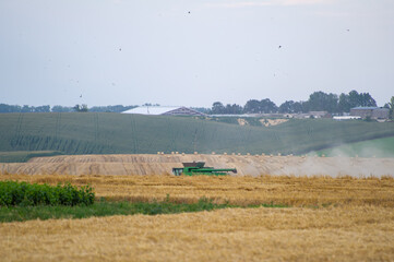 A green combine harvester works across a golden field dotted with hay bales, set against rolling...
