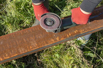 Hands of a worker in safety gloves use an angle grinder to remove rust from a metal beam outdoors....