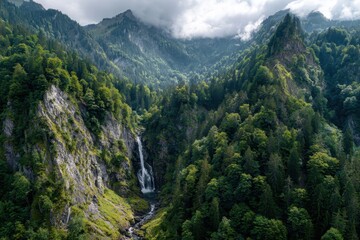 Obraz premium Waterfall cascades through lush forested mountains under a cloudy sky in a serene natural landscape