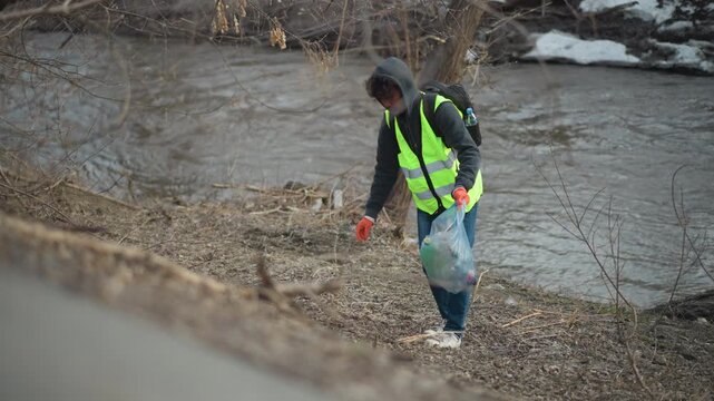 Volunteer wearing reflective vest and gloves collects litter along riverbank, placing trash into blue plastic bag during community cleanup to protect environment and improve natural habitat