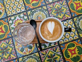 Top view latte art coffee cup and a glass of water on a colorful mosaic tiles background. cappuccino with a heart of foam.