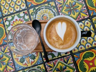 Top view latte art coffee cup and a glass of water on a colorful mosaic tiles background....