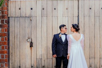 Elegant bride and groom share a tender gaze in front of a large wooden gate, creating a romantic...