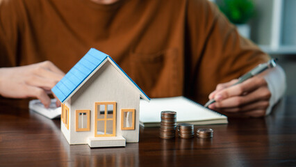 Real estate investment and mortgage calculation concept with businessman using calculator, coins stacked on desk, laptop, and small house model, representing property finance, budget planning, and hom