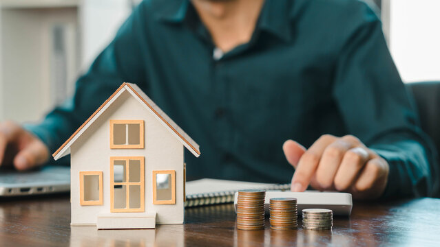 Real estate investment and mortgage calculation concept with businessman using calculator, coins stacked on desk, laptop, and small house model, representing property finance, budget planning, and hom