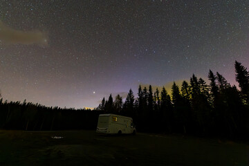 A motorhome parked under a starry sky with the northern lights visible in the forests of Norway.