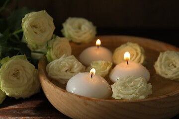 Burning candles, water and rose flowers in bowl on wooden table, closeup